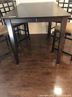Tall wood bar table with dark finish, four square legs, rectangular top, photographed with chairs around it on wooden floor.