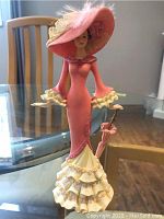 Full view of the pink-dressed figurine showing ruffled tiers, large hat with lace and flowers, and a parasol at her side, standing on a glass table.