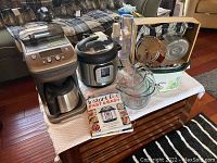 Wide view of all kitchen items on white cloth on wooden coffee table in living room, showing coffee maker, Instant Pot, cookbooks, Pyrex measuring cups, Rival Crockpot and food processor box.