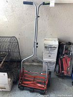 Full view of the manual push lawnmower showing tall silver handle with black grips and red-orange base with wheels.