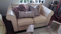 Wide view of cream leather loveseat with four decorative cushions on it, showing overall condition and style.