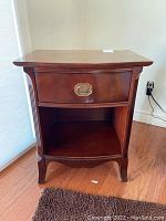 Front view of the wooden night table showing the drawer and bottom shelf, placed on wood floor with a rug in front.