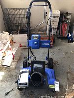 Front view of SnowJoe cordless snow blower with charger and battery base placed on floor.