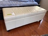 Full view of leather storage bench, showing off-white leather upholstery and wood legs on hardwood floor.
