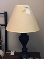 Photo of one lamp with beige shade and black base, labeled with tag showing dimensions and lot number.
