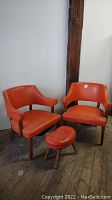 Two orange Mid Century Modern chairs and one matching foot stool with wooden legs, placed on a rustic wooden floor against a light grey wall