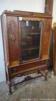 Front view of the antique burled wood china cabinet showing central glass door, side wood panels with carvings, lower drawer, and turned legs.