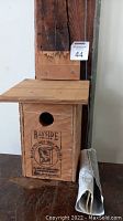 Front view of handmade wooden birdhouse branded with Bayside Prison logo and papers beside it.