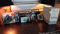 Photo showing the entire assortment on a black surface under lamps including boxed Sony phone, computer tower, camera, radios, calculators and office devices.