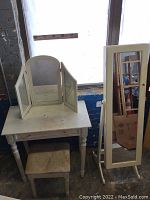 White wooden vanity table with tri-fold mirror, matching stool beside a tall standing jewelry armoire with mirror and lock, all against a windowed brick wall.