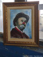 Framed clown portrait artwork depicting a sad clown in dark clothing and a brown hat with a red nose, set against a blue sky background, hanging by a window.