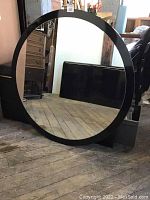 Photo of large round mirror with black frame standing on wooden floor leaning against wall, showing the size and condition of the mirror.