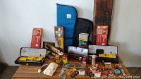 Overview photo showing multiple boxed and unpacked rifle cleaning kits, gun oils, patches, brushes, and two rifle bags against a white background.