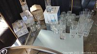 Clear assortment of glass drinkware arranged on a glass table, including multiple tall and short tumblers, mason jars, small shot glasses, and stemmed wine glasses. Some boxed glasses visible in background. Needs cleaning.