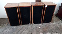 Four vintage floor standing speakers arranged side by side showing black fabric fronts and wooden top and side frames.