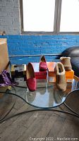 Four pairs of women's shoes arranged on glass table in front of blue brick wall. Includes red loafers, tan boots, purple ballet flats, and boxed shoes.