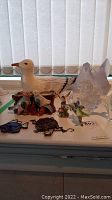 Wide shot of ceramic seagull, glass rooster dish, glass lion, ceramic birds on branch and stained glass fish and turtle decorations arranged on white surface near window with blinds.