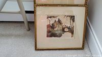 Three framed prints on floor, show artwork of village with bridge and houses, set against carpet. Frames have gold sign of wear