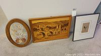 Three framed wall art pieces leaning against a wall: oval pressed flower art, rectangular wood inlay bridge scene, and smaller signed artwork.