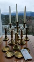 Full set of six brass candlesticks including three single pieces, one three-arm candelabra, and six round coasters arranged on a table in front of a window.