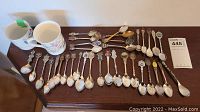Full layout of 33 souvenir spoons and two ceramic cups on wooden surface, spoons have various decorative emblems and handles.