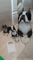 Four Boston Terrier dog figurines placed on the floor, showing size variation with the largest figurine measuring about 16.5x7x8.5 inches as noted on a card in front.