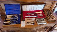 Photo showing entire lot with fish set, Louis Rose carving set, nut crackers, and boxed cutlery sets on wooden furniture.