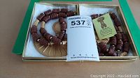 Full view of the necklace displayed in a box showing large brown wooden cylindrical beads with gold-tone spacers and large semicircular pendant. Informational tag included.