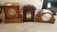 Three wooden mantle clocks with different shapes and finishes shown side by side on carpeted floor.