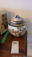 Front view of the porcelain pot showing detailed hand-painted Geisha figures, blue and gold detailed lid and rim, three small feet.