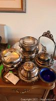 Photo showing multiple silverplate covered dishes including a cobalt blue ceramic insert dish, a silverplate wine bottle holder and a silver kettle with lid on a wooden drawer.