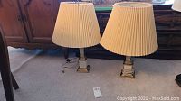 Full view of the pair of matching table lamps with pleated cream shades on carpeted floor in front of wooden furniture.