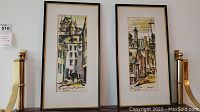 Two framed watercolor paintings shown hanging side by side on a wall, depicting urban architectural scenes in muted tones.
