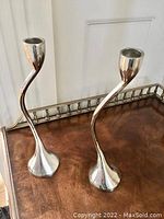 Photo shows both identical silver-tone twisted metal candle holders with flared round bases and cup tops for candles, placed on wooden surface with white wall background.