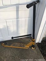 Full side view of the gold and black kick scooter leaning against a door, showing the deck, handlebars, and wheels.