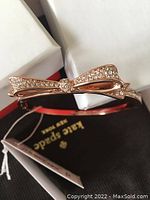 Rose gold tone bangle bracelet with a crystal-studded bow on top, placed near a Kate Spade New York pouch and box.