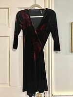 Front view of black velvet wrap dress with red floral pattern, 3/4 sleeves, and waist tie hanging on hanger.