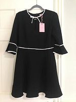 Full front view of the black Ted Baker dress with white piping on neckline, sleeves, and waist, hanging on a hanger against a white door.