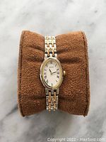 Front view of Seiko ladies watch showing oval white dial with Roman numerals and two-tone silver and gold linked band resting on a brown cushion.