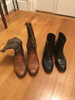 Pair of boots on wooden floor showing brown Tony Lama cowboy boots with patterned stitching and pointed toes, and black Florsheim leather boots with block heels and zippers.