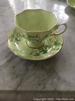 Single bone china cup with matching saucer in pale green with floral vine pattern and gold trim placed on marble surface.