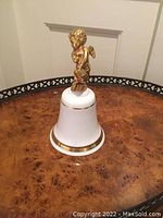 White porcelain bell with gold trim and gold cherub figure on top, shown on a wooden table with decorative edge.