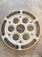 Top-down photo of the white ceramic Seder plate with blue, gold, and black ornamental patterns and seven distinct round sections for Passover ceremonial items.