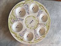 Top-down image of round ceramic Seder plate with seven indentations and decorative grapevine and olive branch artwork. Hebrew inscriptions visible in each indentation and center.