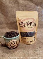 Bag of Guatemala Elpida organic coffee beans next to a ceramic mug filled with coffee beans on burlap background.