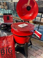 Kamado Joe Kettle Joe grill assembled with lid open on wheeled cart; cooking grate installed; instruction manual on grate; red Kamado Joe Big Block lump charcoal bag, fire starter box and grill cover packaging visible