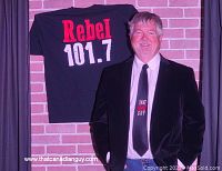 Glen Foster standing in front of a brick wall with a Rebel 101.7 T-shirt hung behind him, wearing suit and tie reading “That Canadian Guy”
