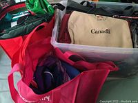 Photo showing various bags including a beige tote bag with "Canada" logo, red and pink bags, green gym bag, and more inside a plastic bin and bags.