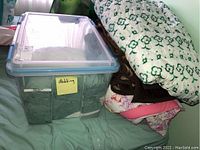 Photo showing a large plastic bin labeled 'Bedding' filled with pillows and shams, with three large blankets piled next to it. Blankets include a floral quilted one, a brown one, and a pink floral fabric item.