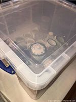 Photo of assorted creamers, lidded sugar bowls, salt and pepper shakers, glass juice glasses, and tin cups inside a clear plastic bin.
